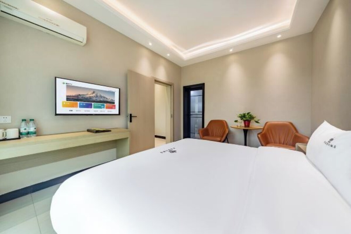 Quarto de hotel com uma cama grande, televisão fixada na parede, uma mesinha com cadeiras e um vaso de planta.