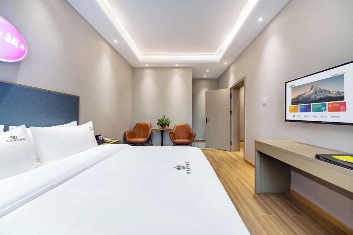 A modern hotel room with a large bed, two armchairs, a small table with a plant, and a TV on the wall.