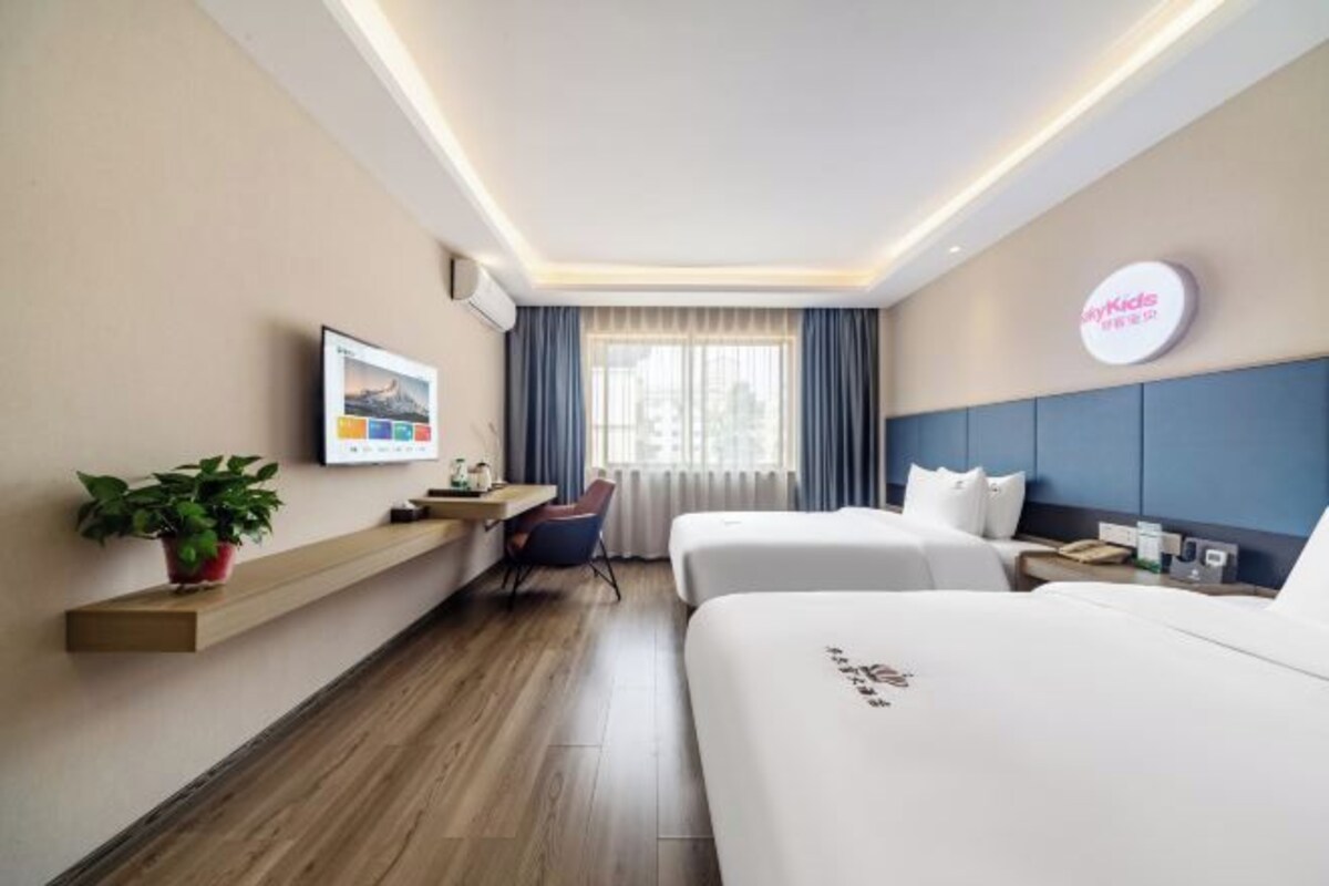 Quarto de hotel com duas camas, uma escrivaninha, uma cadeira, uma planta em vaso e vista para a cidade pela janela.