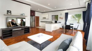 A modern living room with a sofa, coffee table, and entertainment unit. A kitchen with a dining area is visible in the background. - Melbourne Residence (Siem Reap)