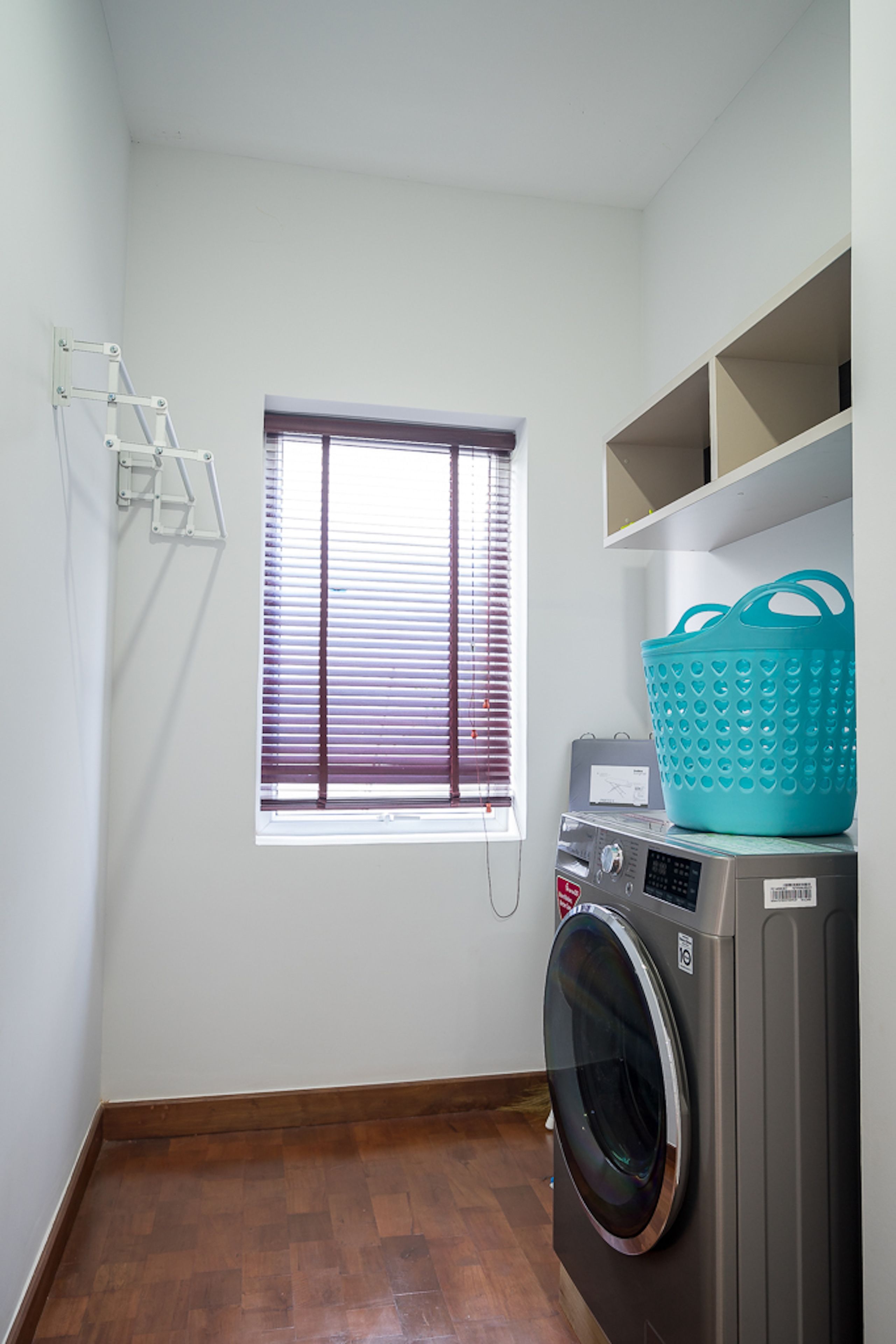 A laundry room with a washing machine, a drying rack, a window with blinds, and a teal basket on top of the machine.