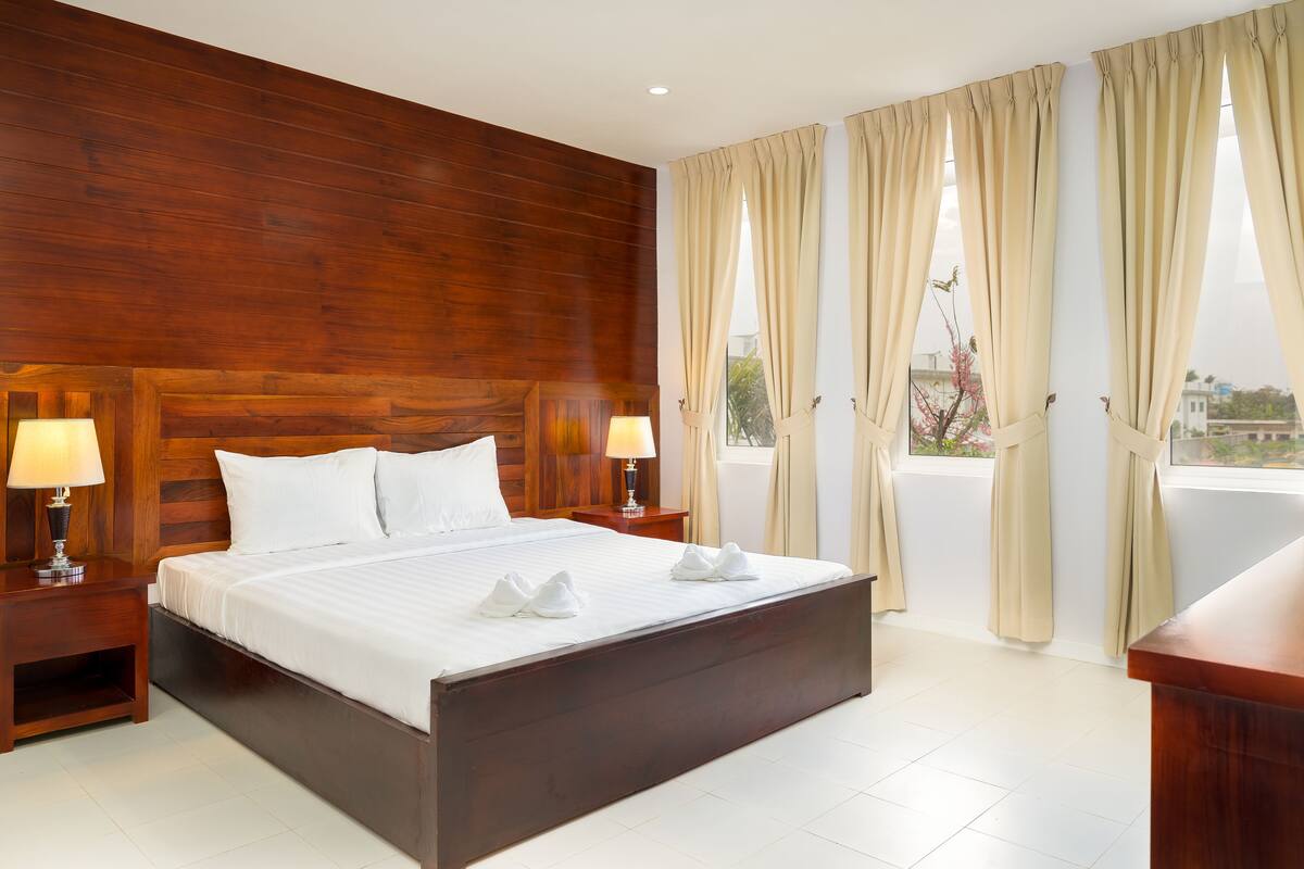 A hotel room with a wooden headboard, a large bed with white linens, two bedside lamps, and beige curtains.
