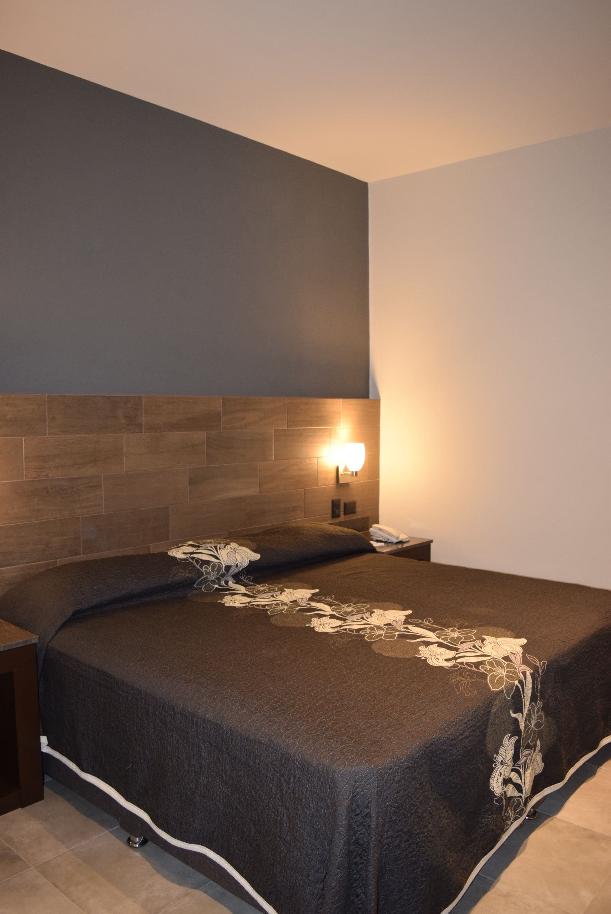 Une chambre d’hôtel moderne avec une tête de lit en bois, une table de chevet avec une lampe et un lit bien fait, recouvert d’une couvre-lit à motifs floraux.