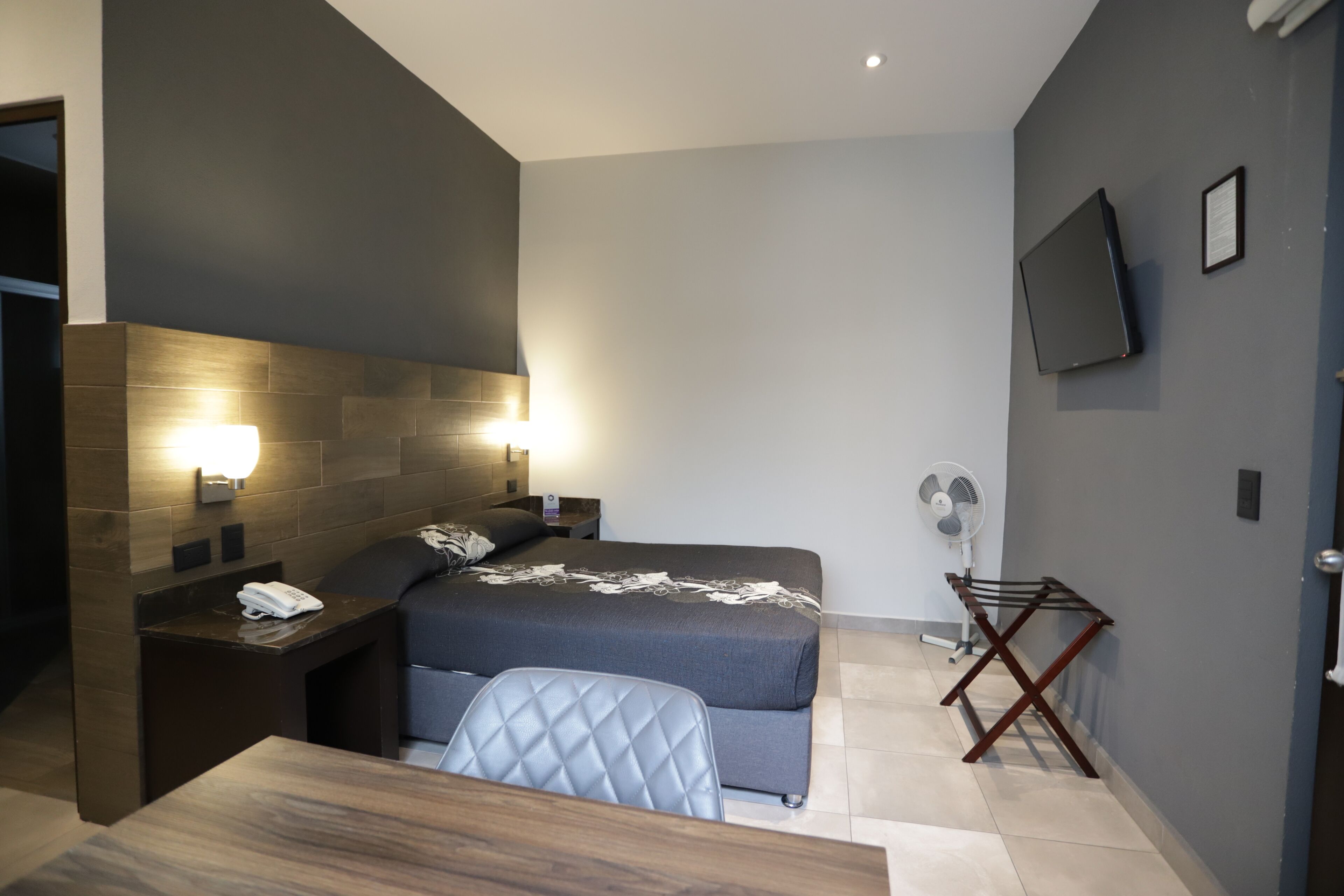A modern hotel room with a bed, bedside tables, a desk, a chair, a TV, and a fan.