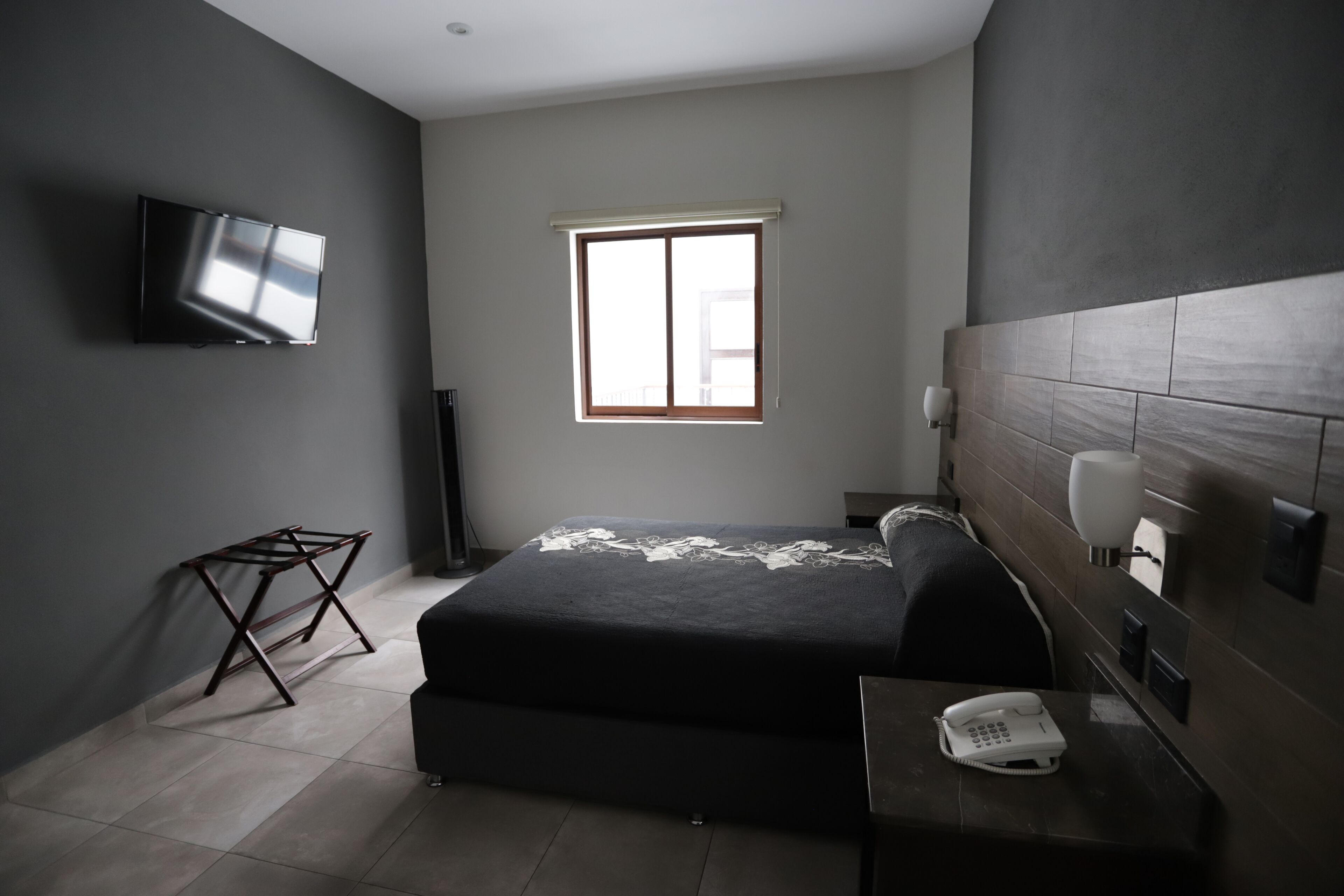 Un dormitorio moderno con cama, televisor montado en la pared, teléfono en la mesita de noche y ventana con persianas.