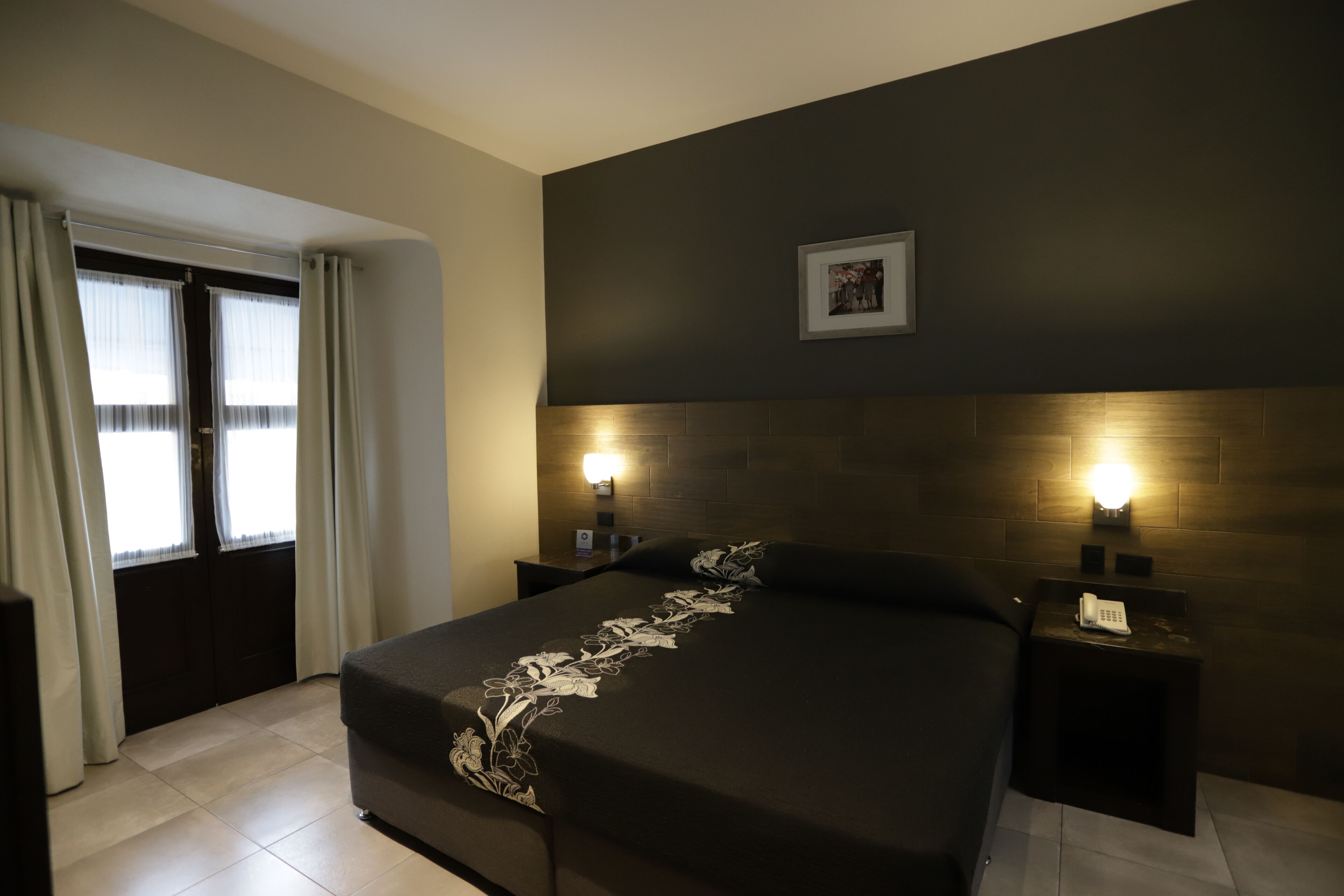 A bedroom with a large bed, two bedside tables with lamps, a framed picture on the wall, and a window with curtains.