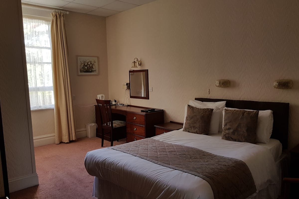 A hotel room with a large bed, a desk, a chair, a dresser, a mirror, and a window with curtains.