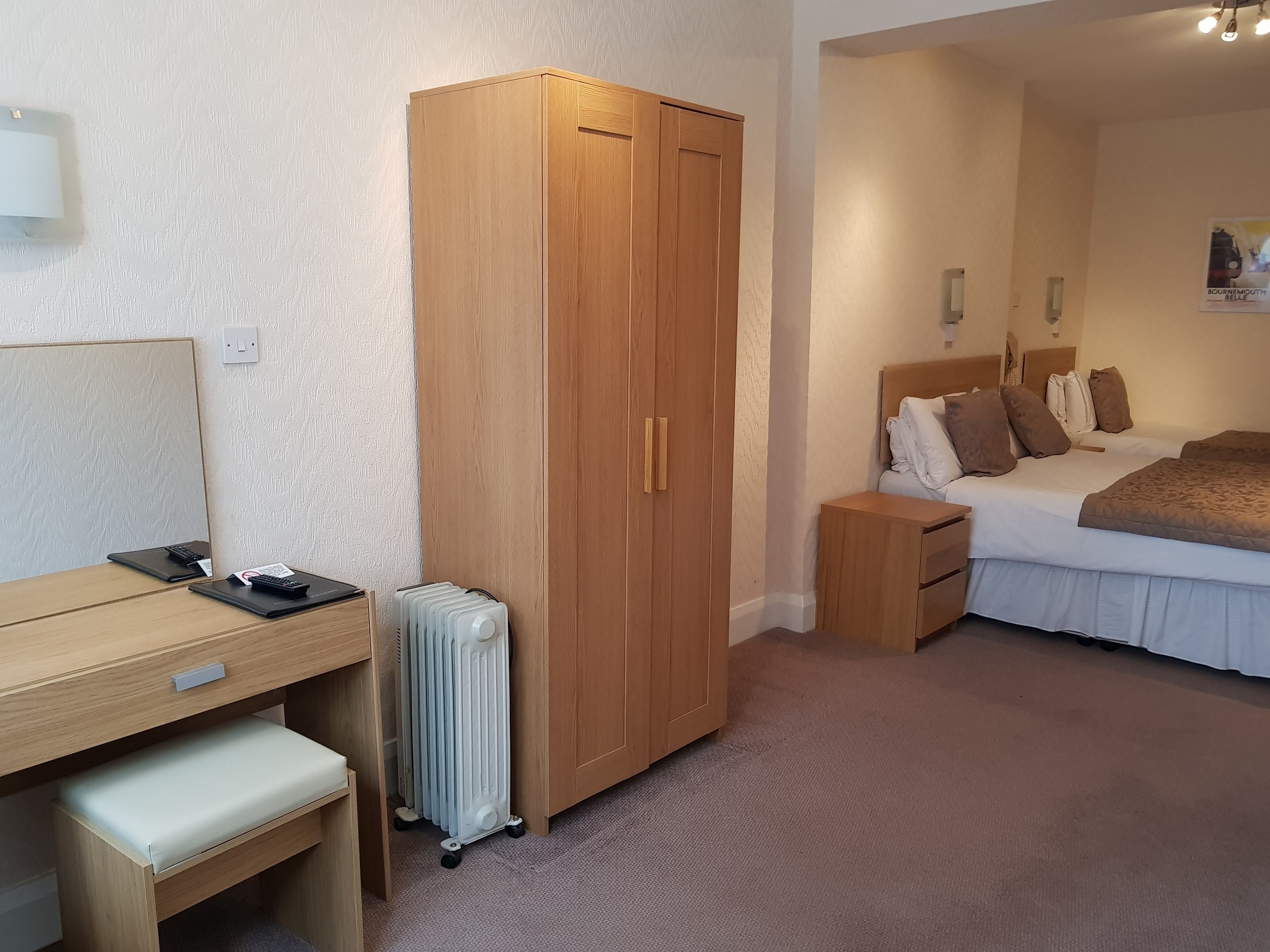 A hotel room with a wooden wardrobe, a bed with pillows, a bedside table, a desk with a chair, and a radiator.