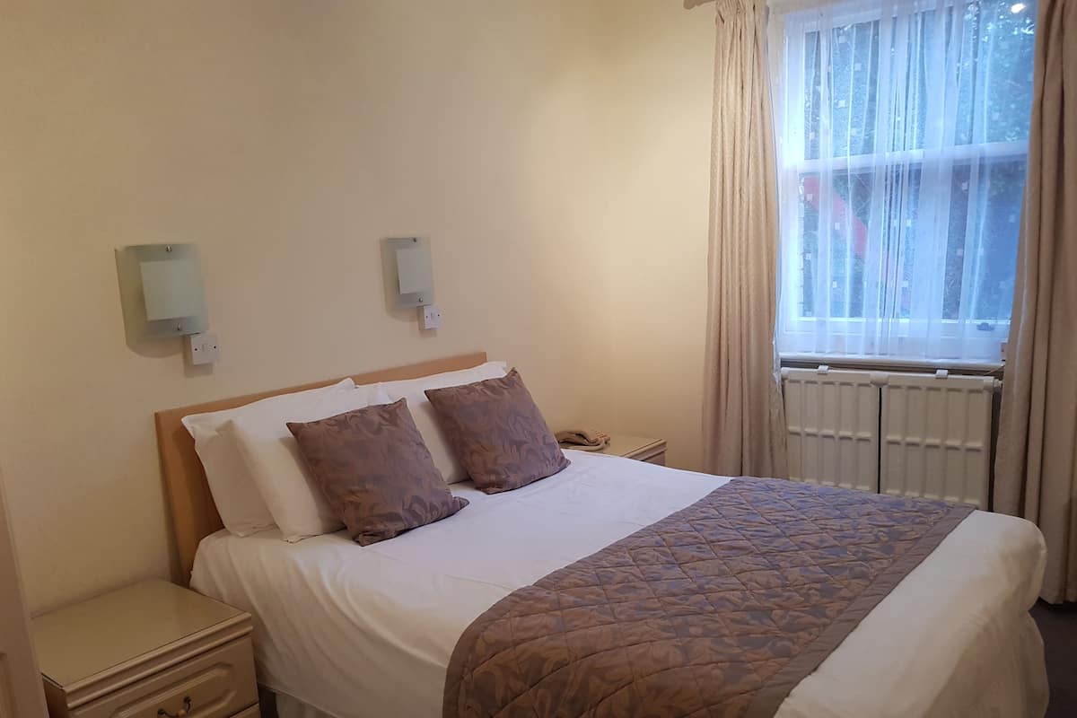 A neatly made bed with a brown quilt, a bedside table with a lamp, and a window with curtains.