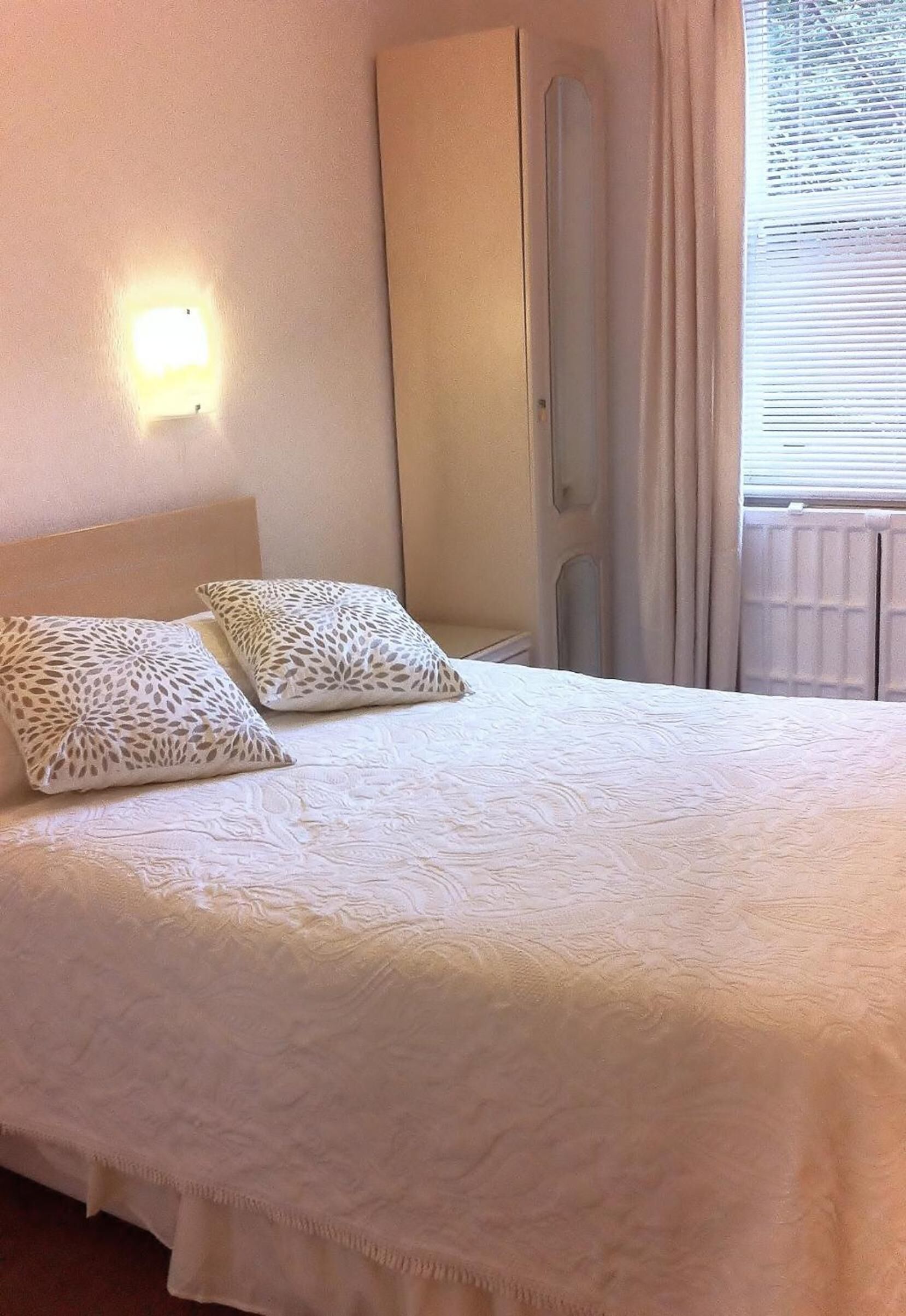 A neatly made bed with a patterned pillow, a wall-mounted light, and a window with curtains.