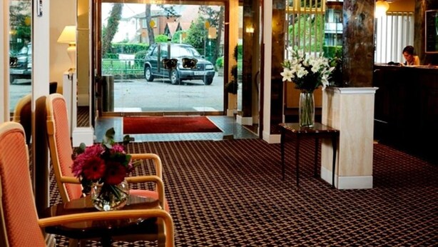 Vestíbulo de hotel con alfombra estampada, una araña y mostrador de recepción, con vista a la calle.