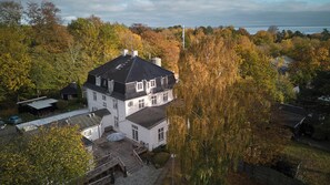 A large, white, multi-story house with a dark roof, surrounded by autumn foliage.