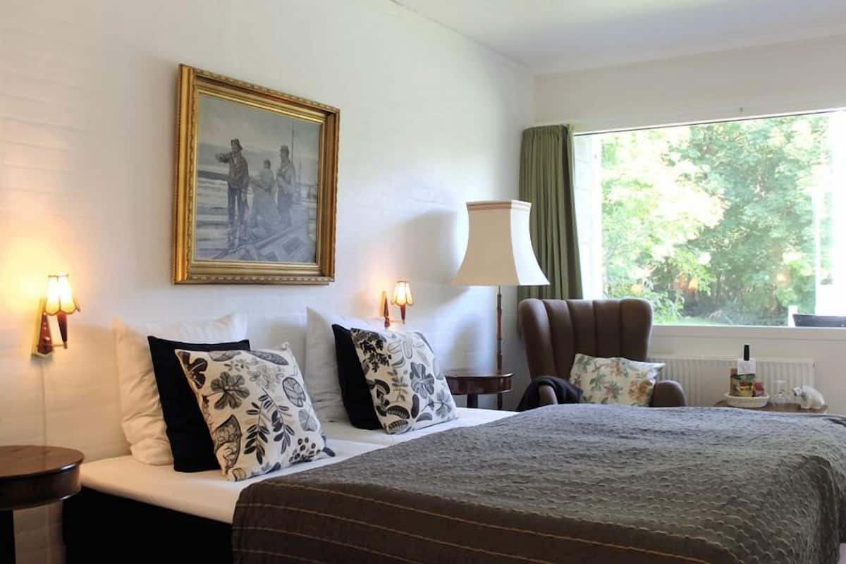 A hotel room with a bed, two bedside lamps, a chair, a window with a view of trees, and a painting on the wall.