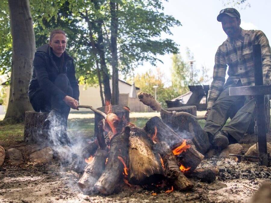 Dos personas cocinando sobre fuego al aire libre, con árboles y un edificio al fondo.