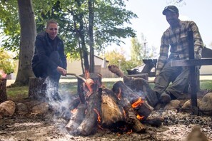 Two people cooking over an open fire in an outdoor setting with trees and a building in the background. - Rorvig Centret (Rørvig)