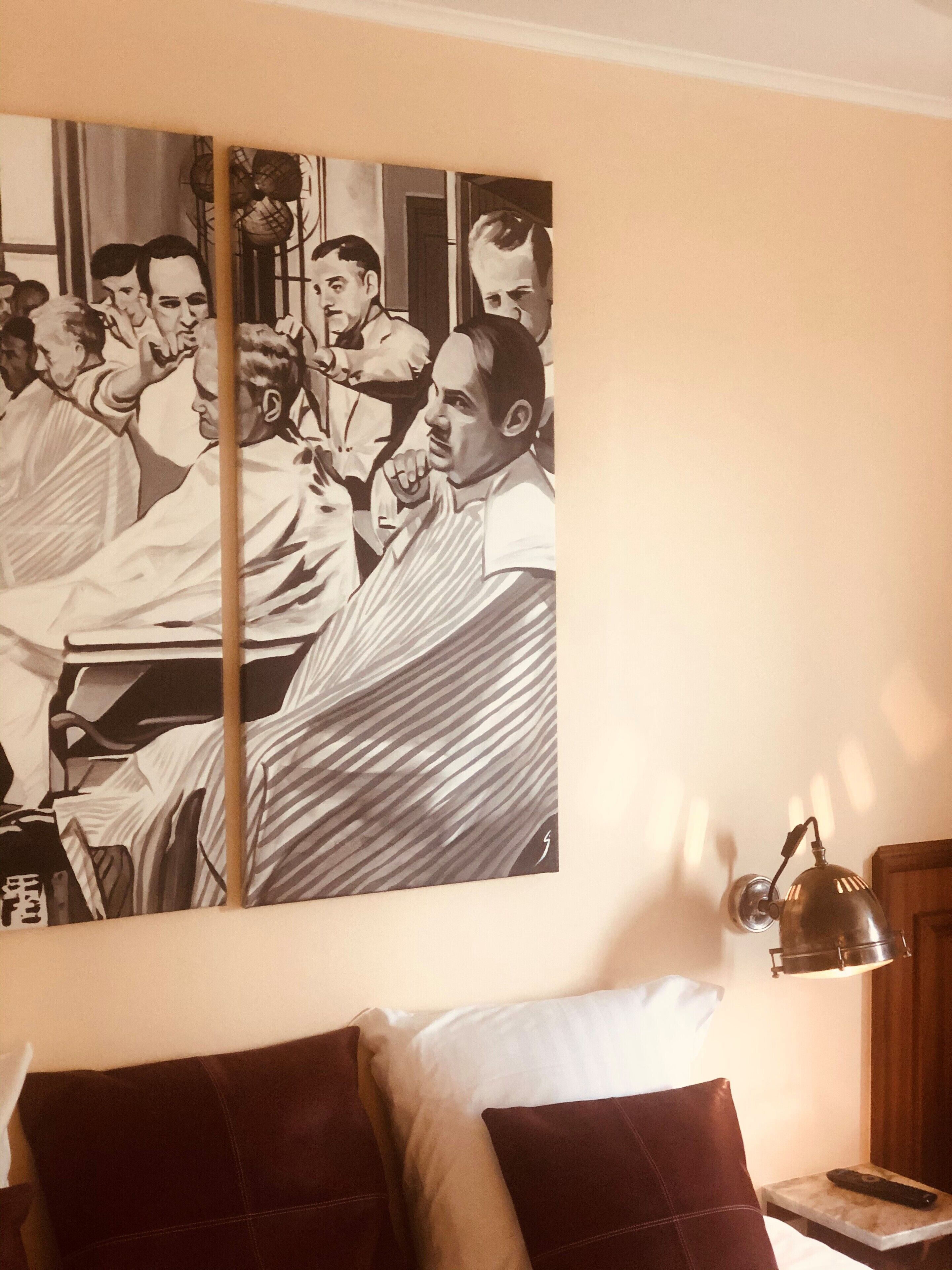 Une chambre d’hôtel avec un grand tableau en noir et blanc au mur, représentant un groupe de personnes réunies. On y trouve un lit avec des oreillers rouge foncé, une table de chevet avec une lampe et une télécommande posée sur la table.