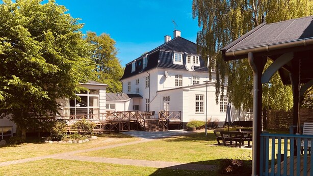 A large white building with a black roof, a covered outdoor seating area, and a paved courtyard. - Rorvig Centret (Rørvig)