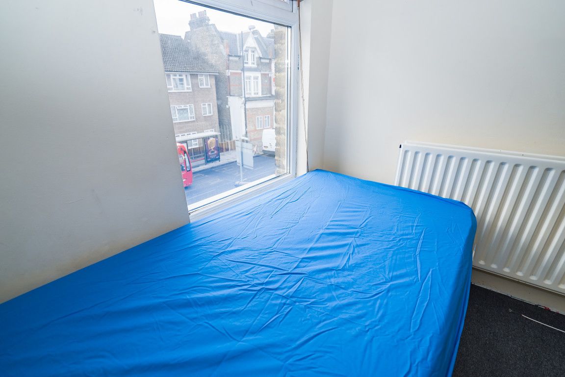 A room with a bed covered in blue sheets, a window with a view of houses, and a white radiator.