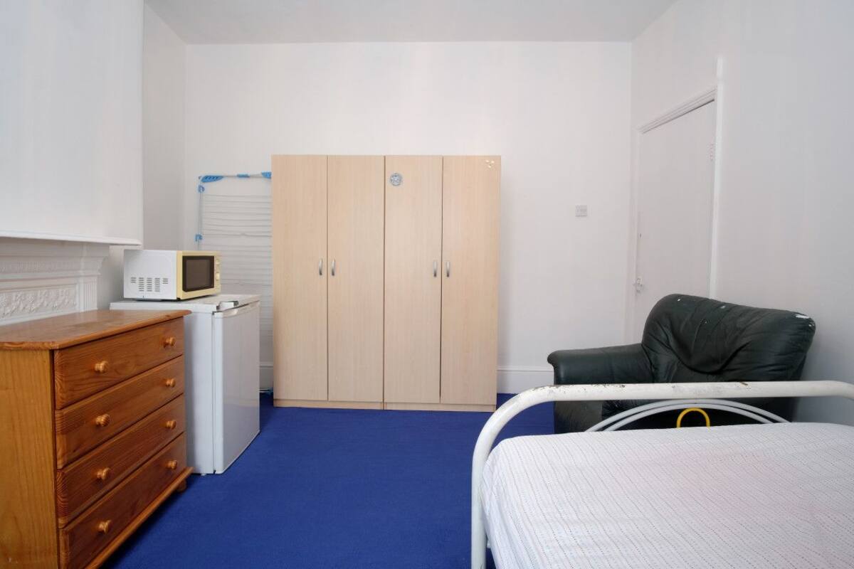 Un dormitorio con una cama, un aparador de madera, un armario blanco, un microondas y una silla negra.
