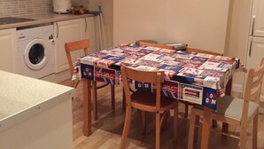 A small kitchen with a table covered in a tablecloth, wooden chairs, a washing machine, and cabinets. - Harley House (London)