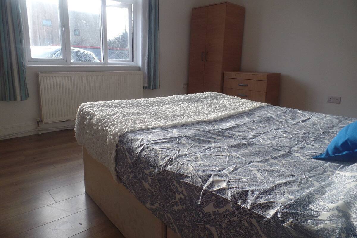 A bedroom with a bed, a radiator, a window with curtains, a wooden wardrobe, and a dresser.