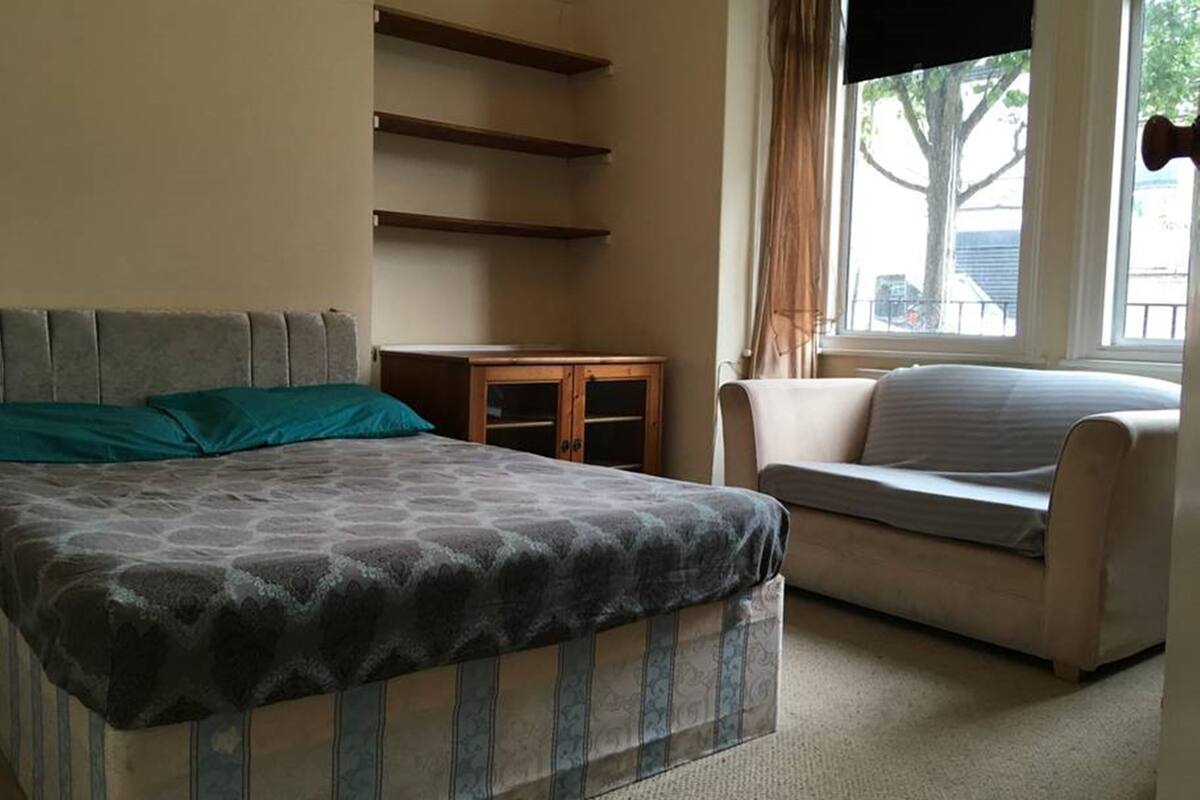 A bedroom with a bed, a small wooden nightstand, a chair, and a window with curtains.