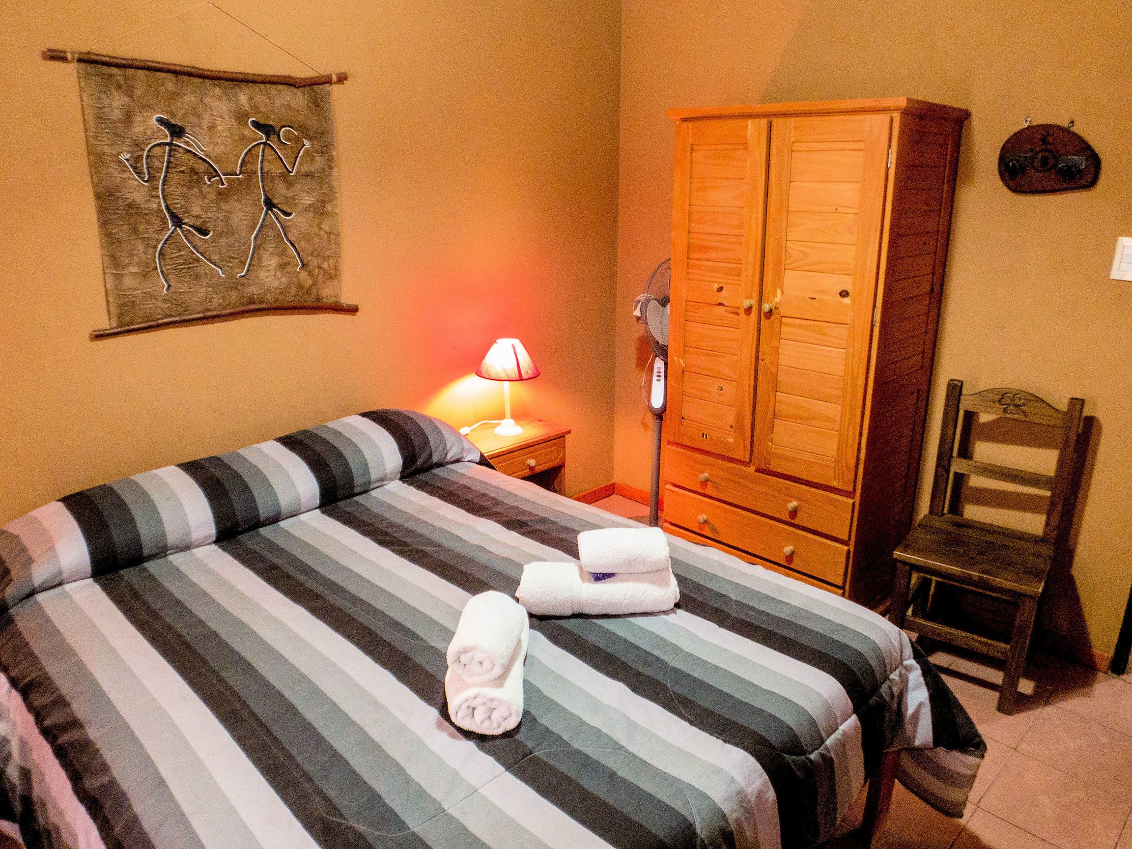 A hotel room with a bed, a wooden wardrobe, a chair, a wall-mounted artwork, and a bedside lamp.