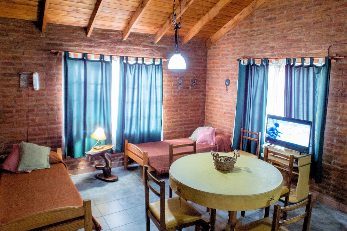 Een gezellige kamer met een bakstenen muur, houten balken in het plafond, een ronde tafel met stoelen, een bank en een televisie.