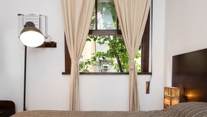 Ein Schlafzimmer mit Bett, einer Wandlampe, einem Fenster mit Vorhängen und Blick ins Grüne.