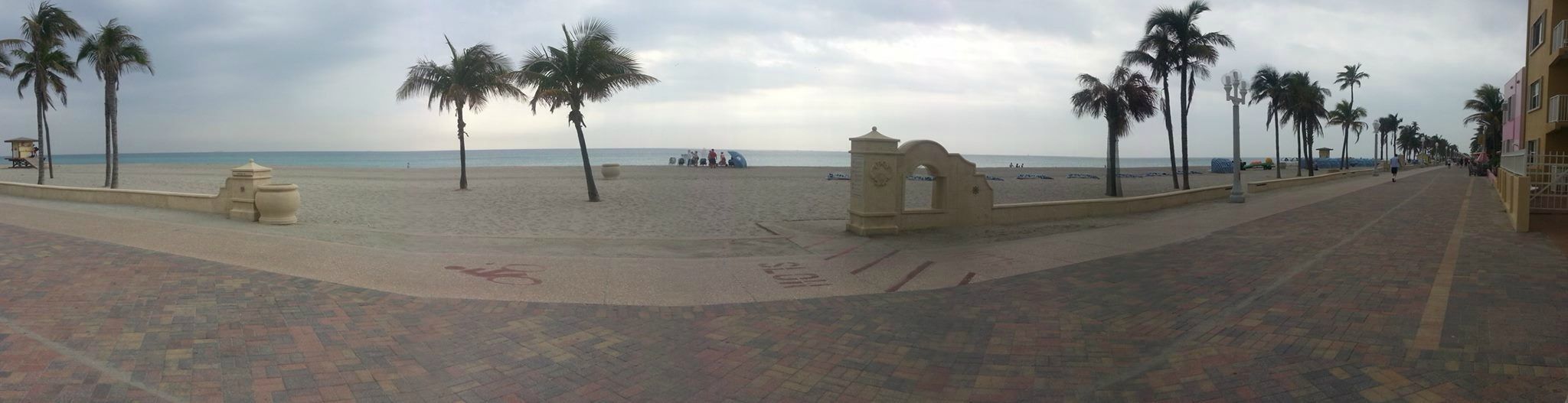 Een promenade aan het strand, met palmbomen, een strand op de achtergrond en een gebouw aan de rechterkant.