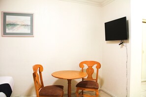 Een kleine kamer met een ronde houten tafel en twee stoelen, een lijst met een afbeelding aan de muur, en een aan de muur bevestigde televisie.