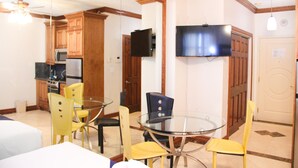 A dining area with a glass table and yellow chairs, wooden cabinets, a TV mounted on the wall, and a ceiling fan.