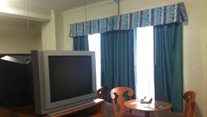 Ein Hotelzimmer mit Fernseher auf einer Holzkommode, einem kleinen Tisch mit Stuhl und einem Bett mit gemustertem Überwurf.