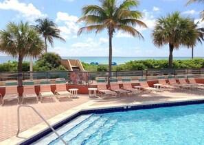 Un espace piscine doté de chaises longues et de palmiers, avec vue sur une plage.