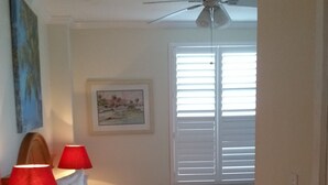 A hotel room with a bed, a desk with a lamp, a ceiling fan, a window with blinds, and a painting on the wall. - Hollywood Beach Resort- One Bedroom Ocean View (Hollywood)