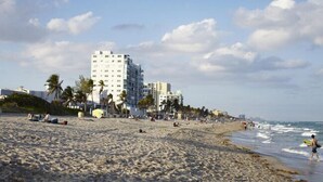 A beach with a large hotel building in the background, palm trees, and people on the sand.