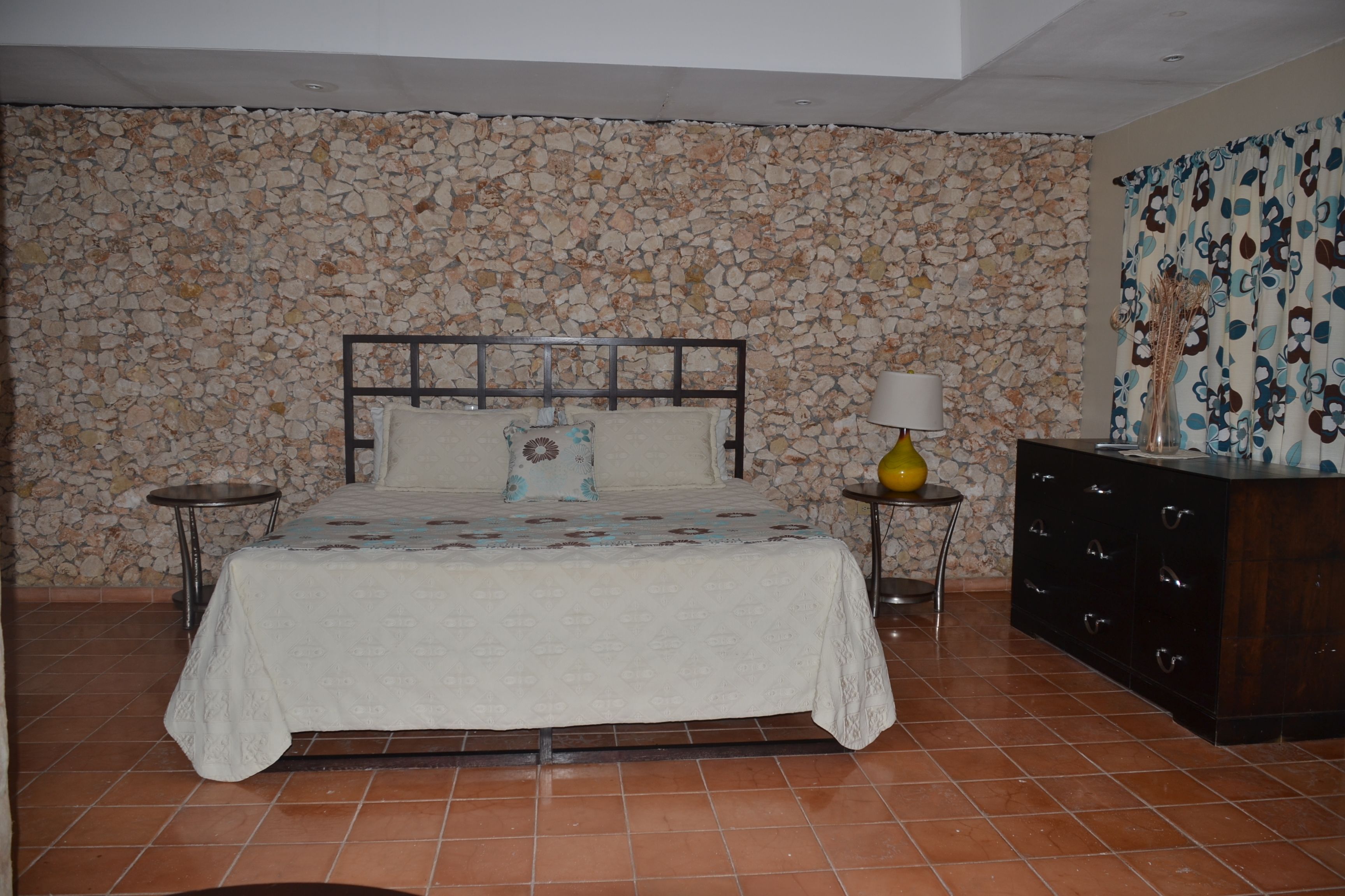 A bedroom with a stone wall, a bed with a patterned bedspread, a nightstand with a lamp, and a dresser.