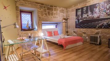 A bedroom with a stone wall, a bed with a red blanket, a glass table with a chair, a window with a ship model, and a cityscape painting.