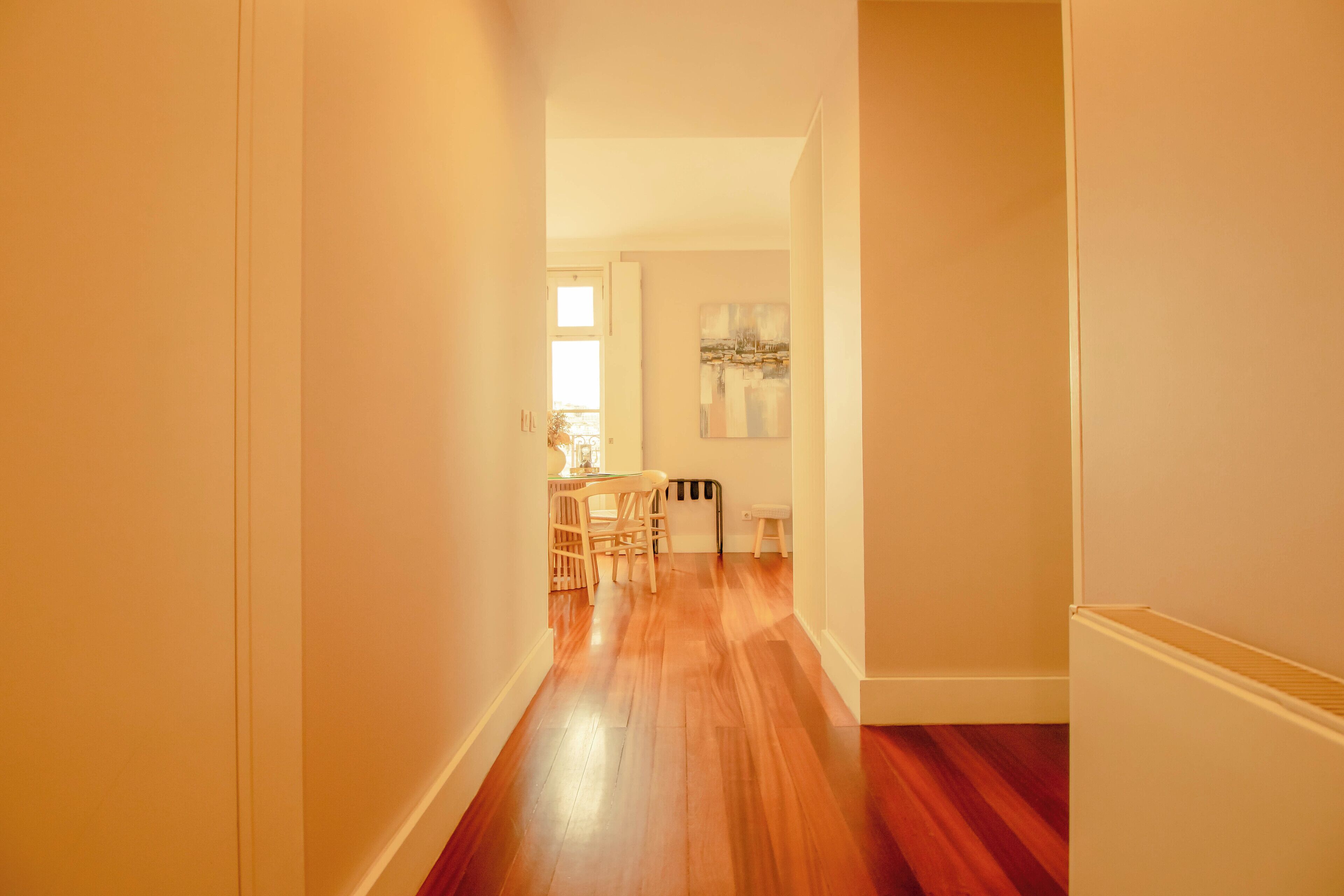 A hallway with wooden flooring, a dining area with a table and chairs, and a painting on the wall.