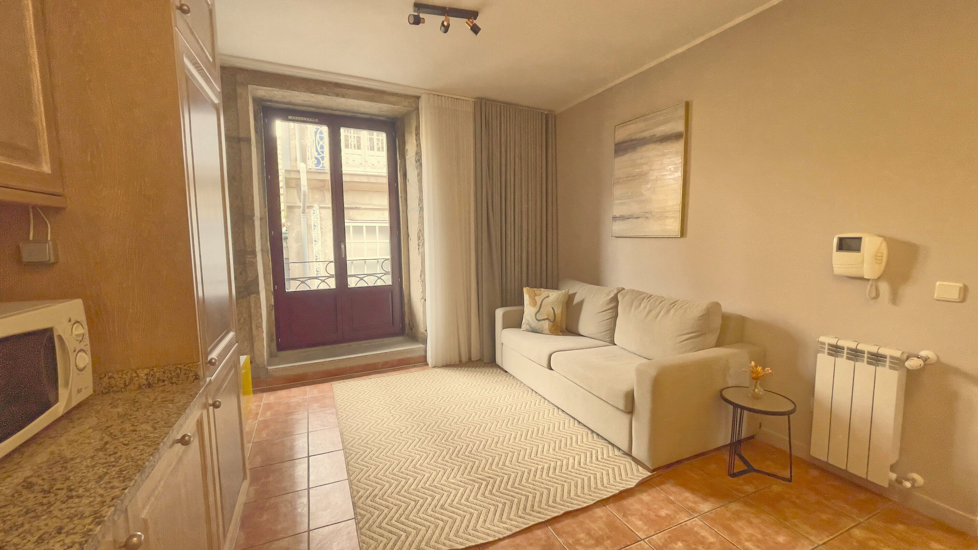 A small living room with a sofa, a small table with a vase, a radiator, and a window with curtains.