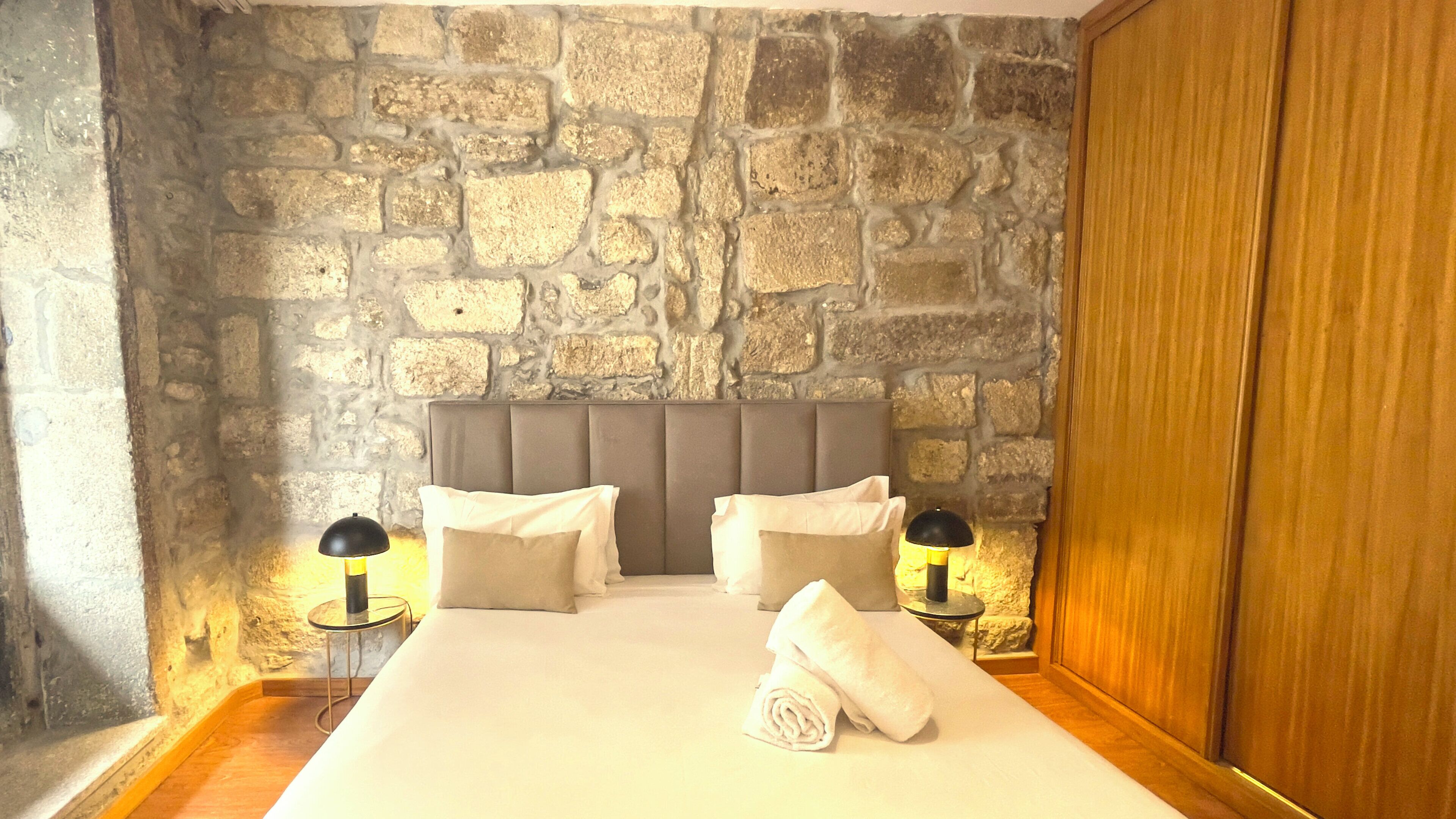 A bedroom with a stone wall, a padded headboard, two bedside tables with lamps, and a neatly made bed with a rolled towel.
