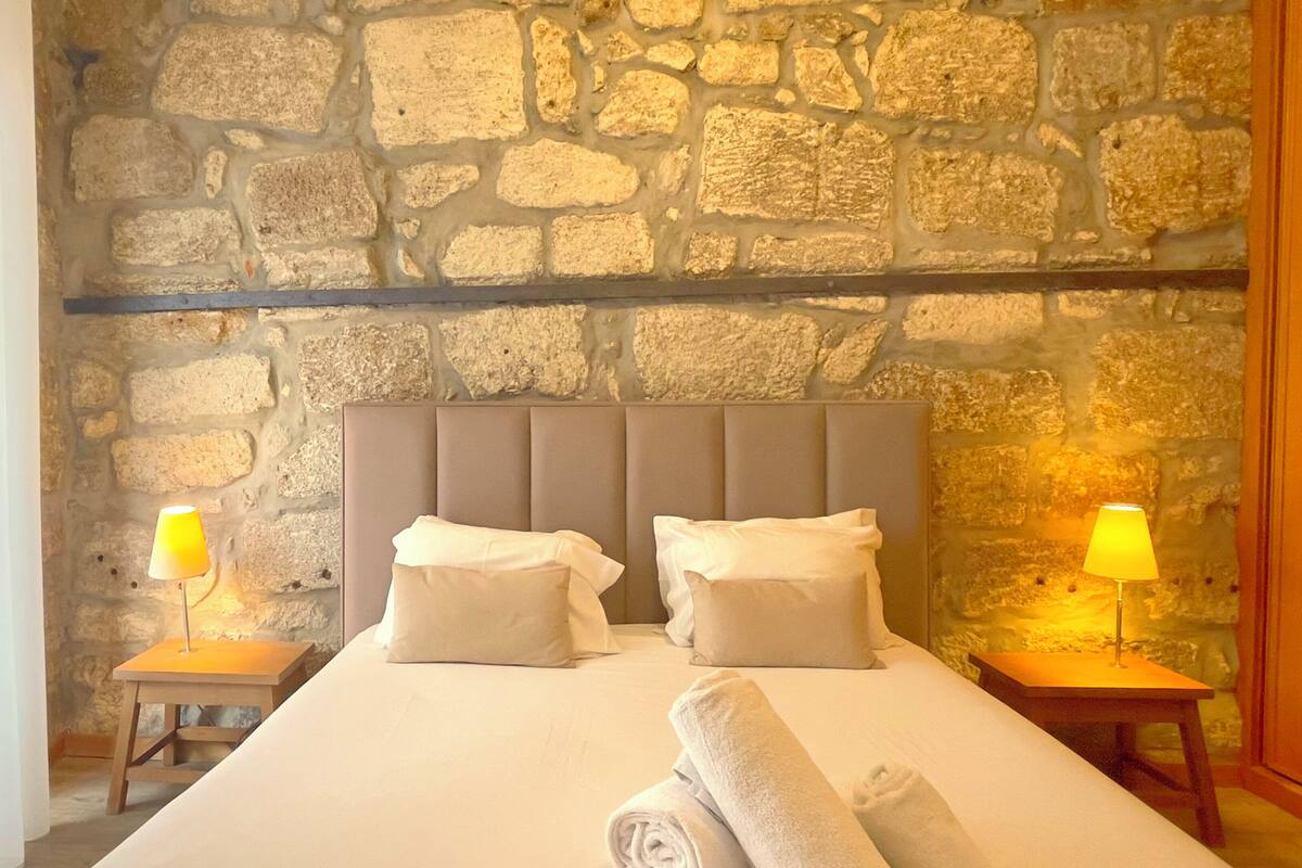 A hotel room with a stone wall, a padded headboard, two bedside tables with lamps, and a neatly made bed with towels.