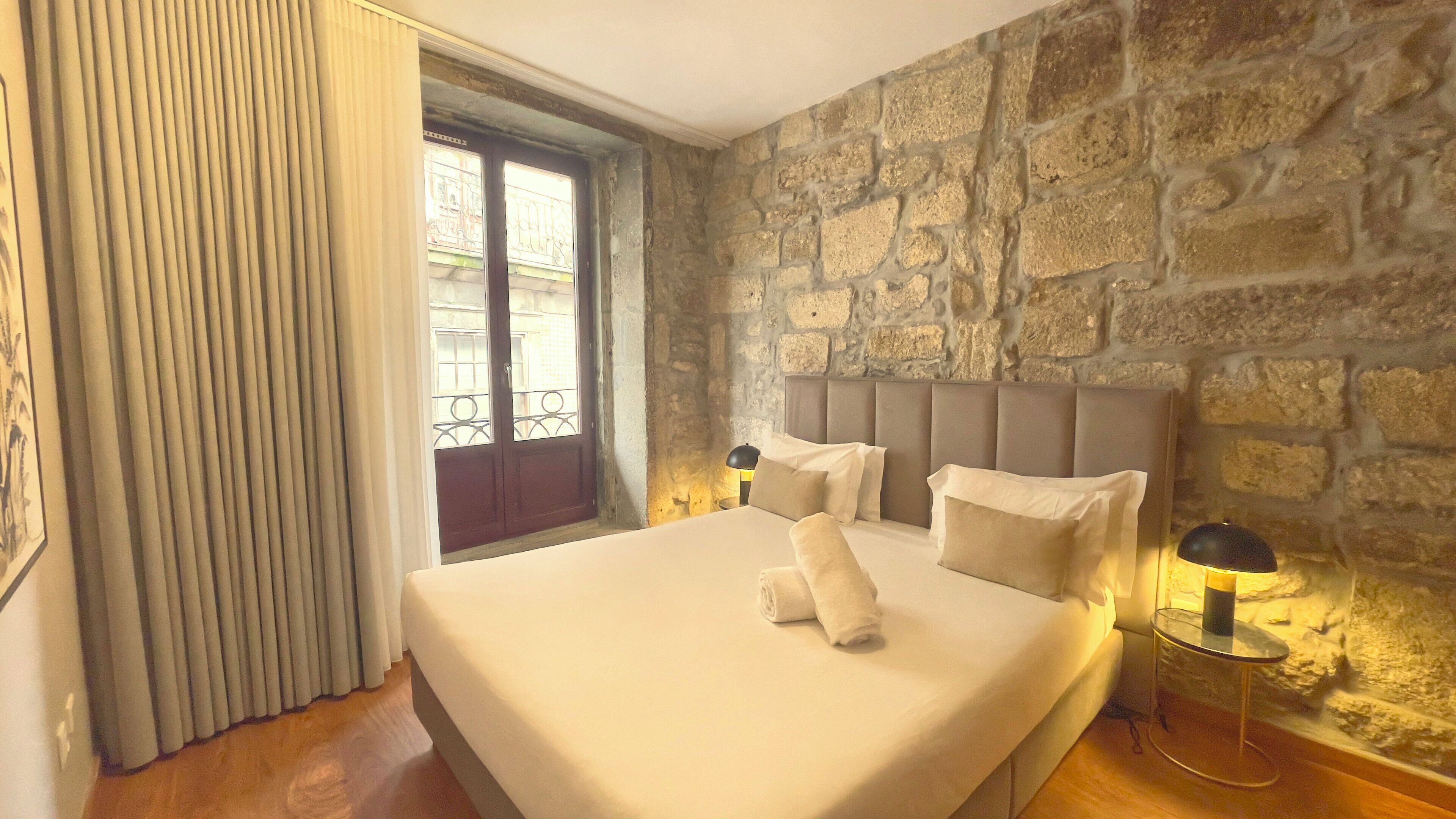 A bedroom with a stone wall, a large window with a view, a bed with pillows, and a bedside table with a lamp.