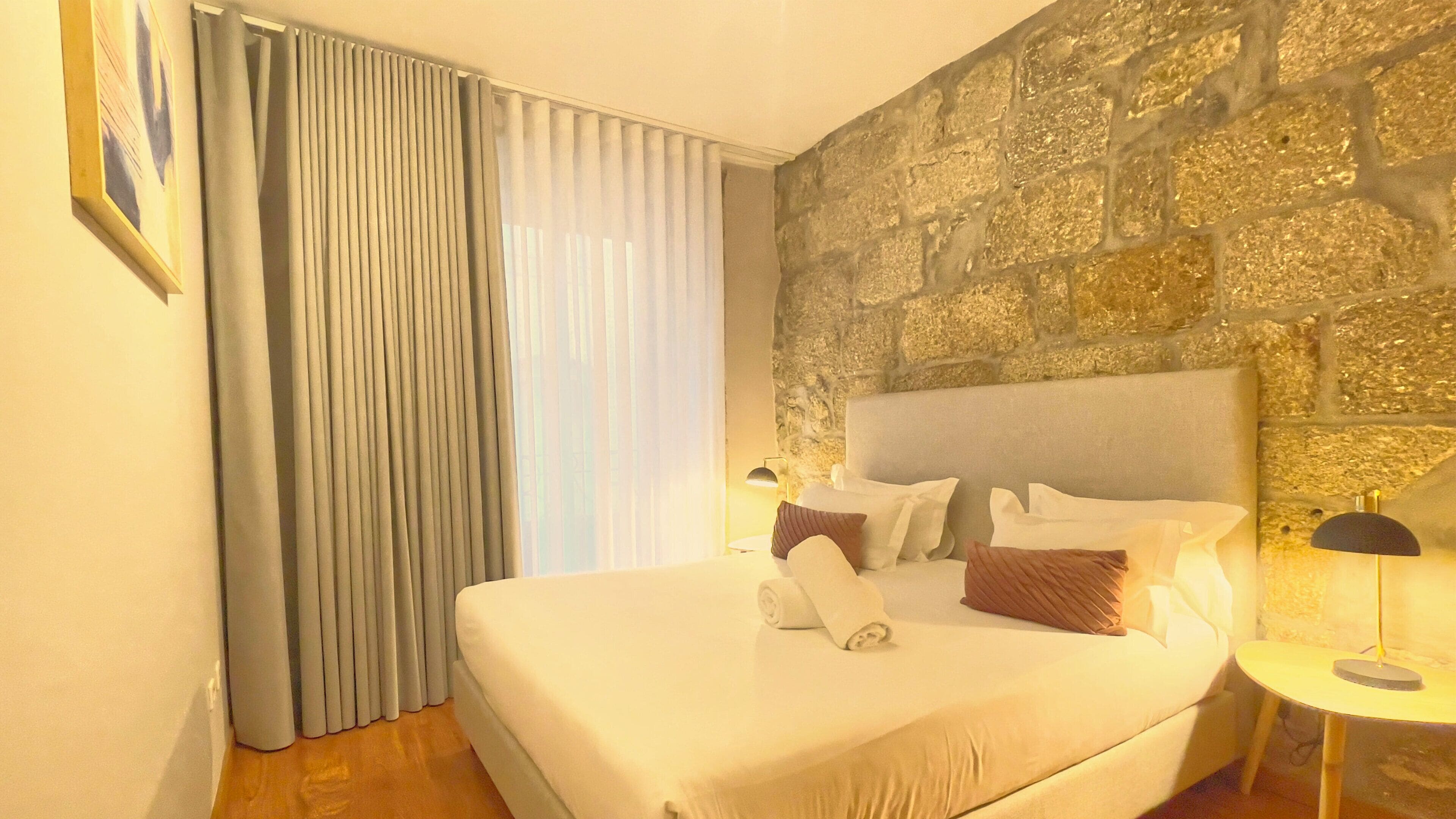 A hotel room with a stone wall, a large bed with white linens, a side table with a lamp, and a window with curtains.