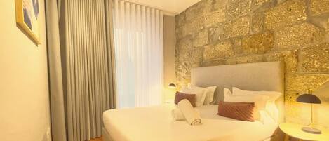 A hotel room with a stone wall, a large bed with white linens, a side table with a lamp, and a window with curtains.