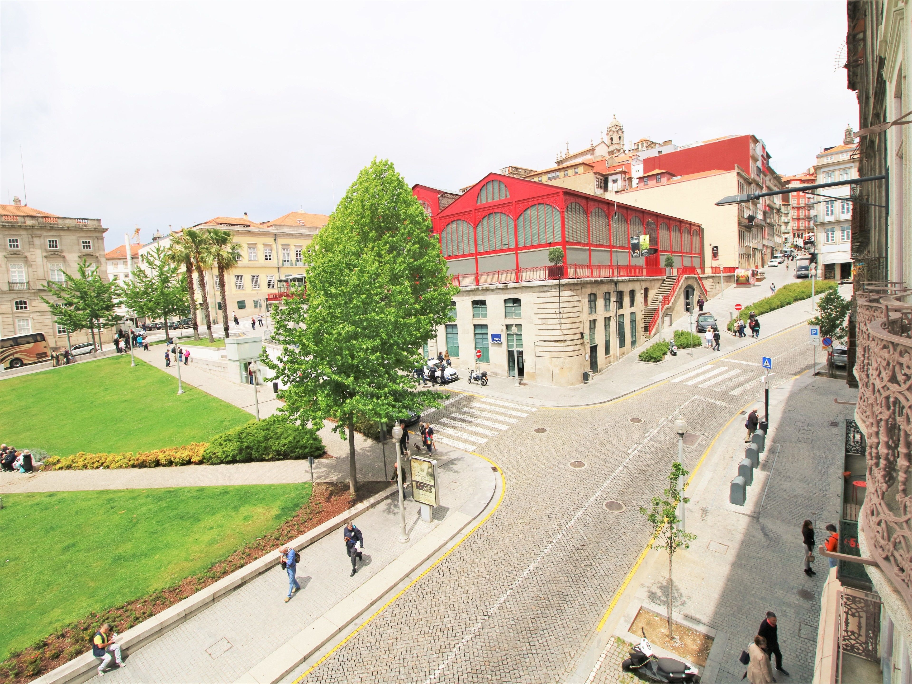 A cobblestone street with pedestrians, a green park, and buildings with red and beige facades.