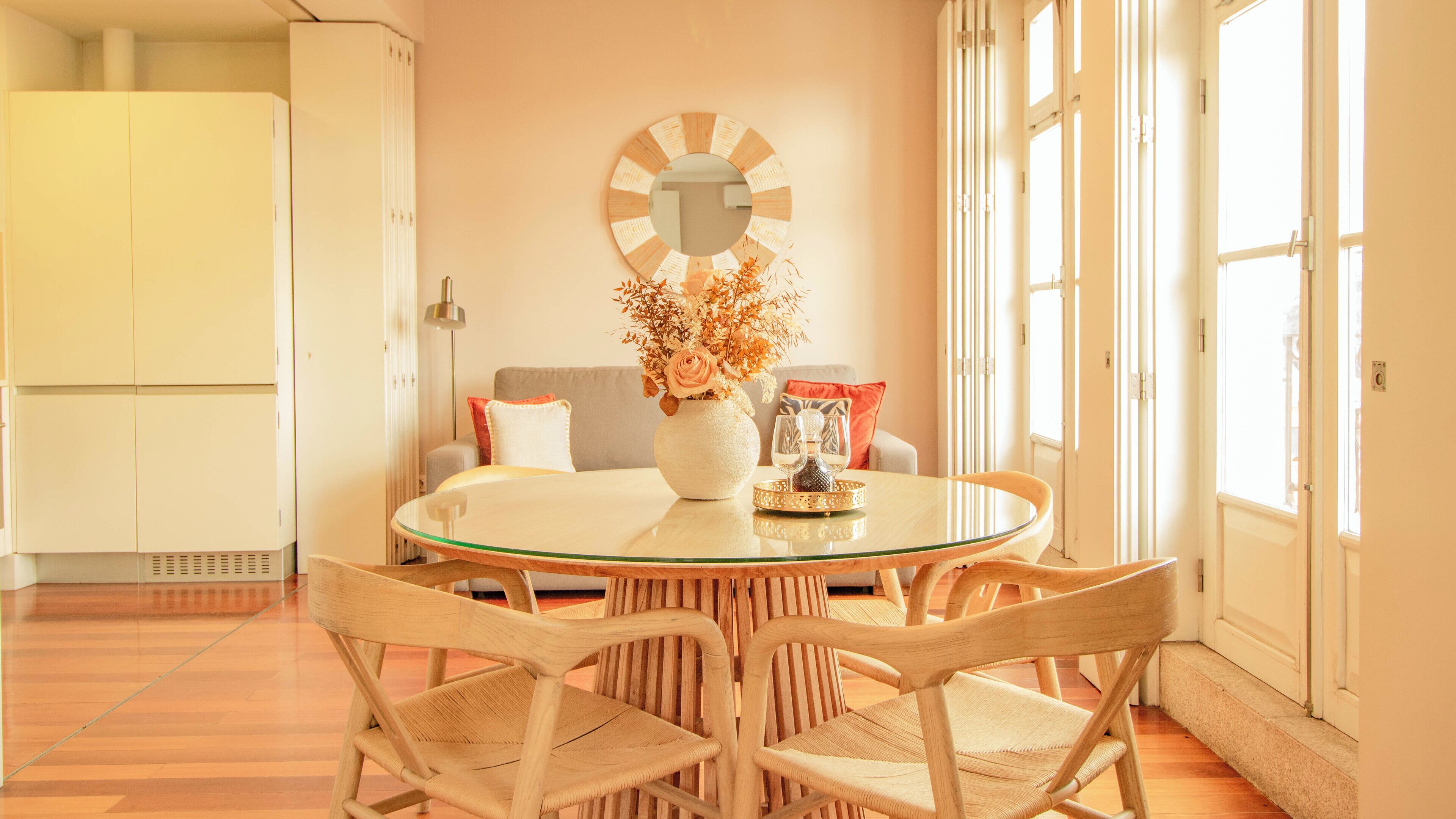 Un coin repas comprenant une table ronde en verre, des chaises en osier, un vase avec des fleurs et un miroir fixé au mur.
