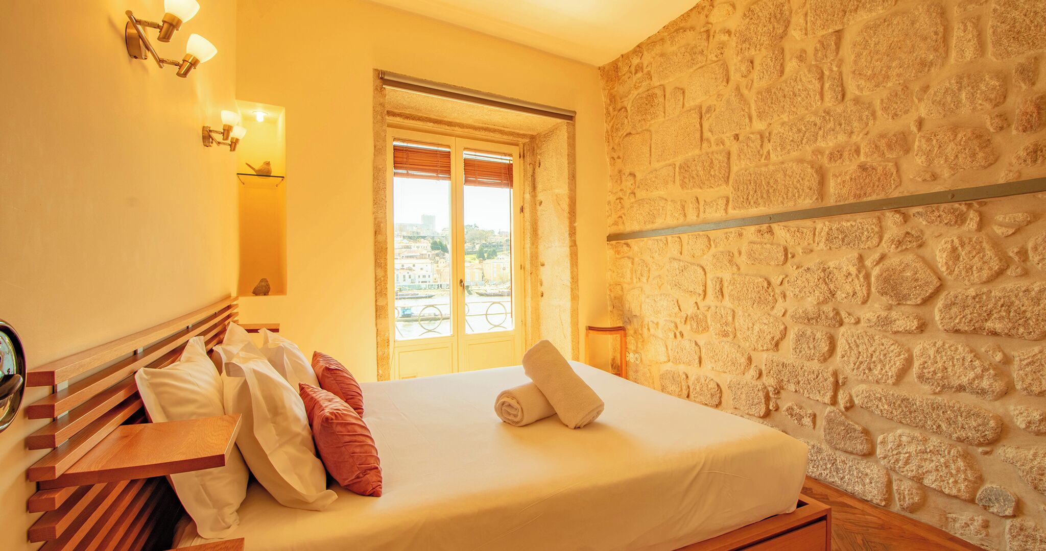 A hotel room with a bed, wooden headboard, stone wall, and a window with a view of buildings.
