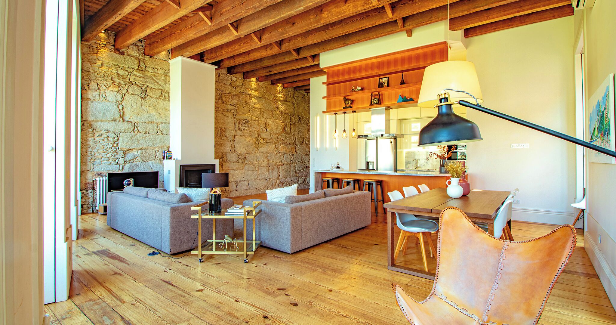 A cozy living room with a stone wall, wooden ceiling beams, a fireplace, a sofa, a coffee table, and a dining area with a table and chairs.