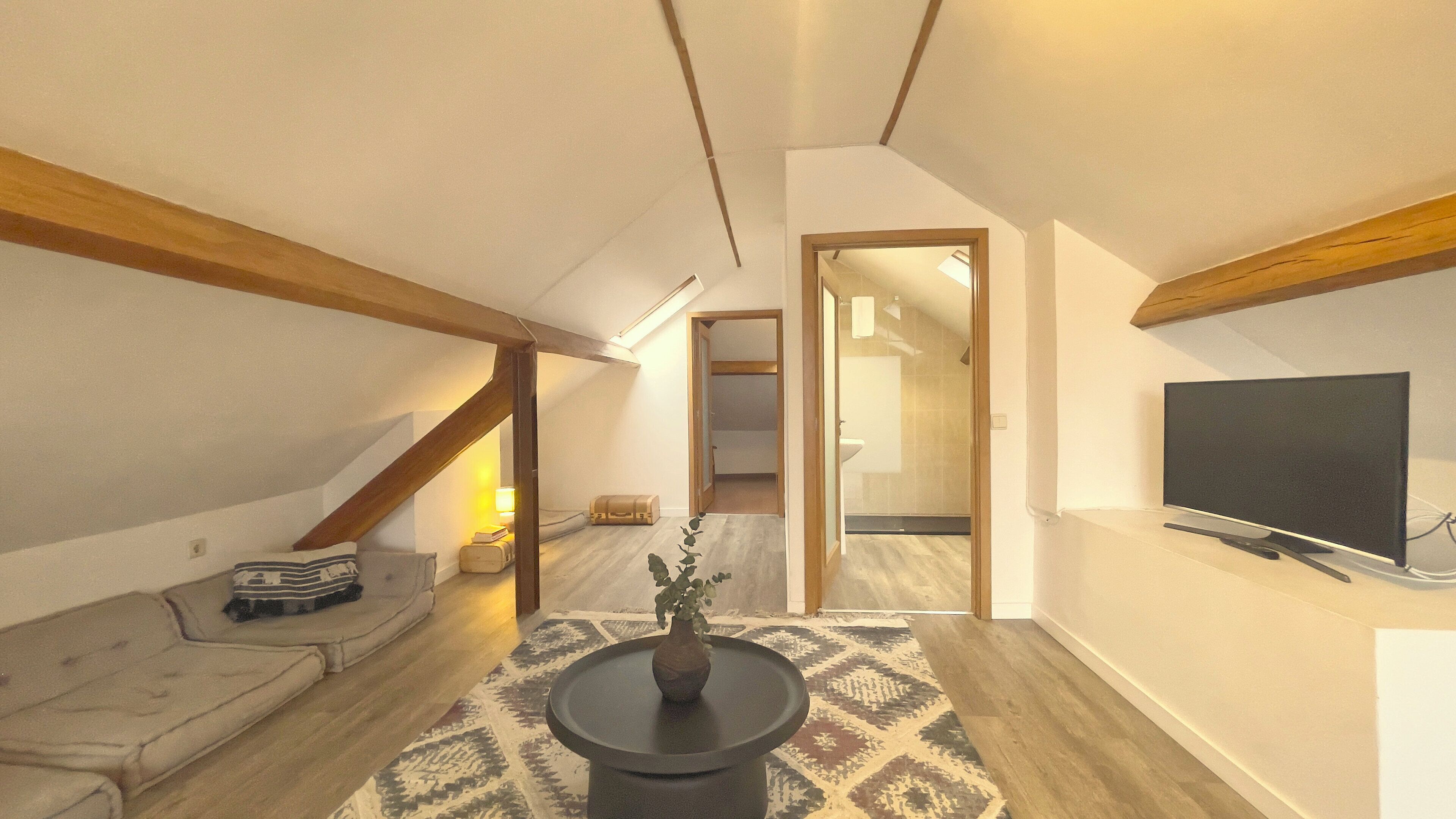 Um apartamento moderno estilo loft com pé direito alto, vigas de madeira aparentes, um sofá, uma mesa de centro, uma televisão e um tapete estampado.